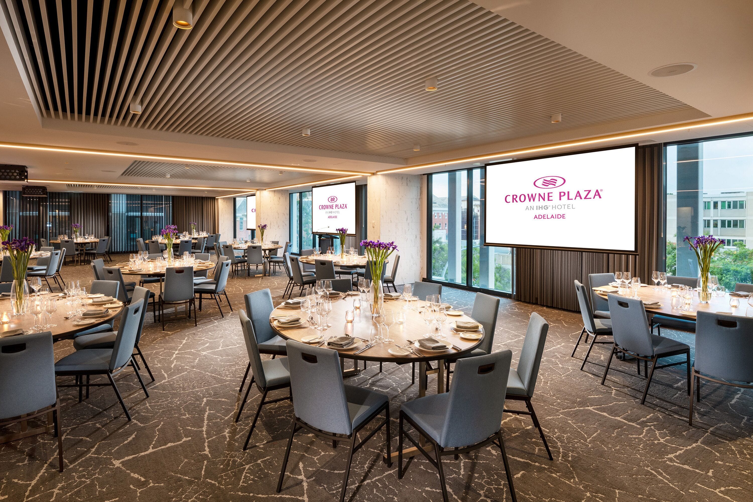 A conference room at Crowne Plaza with multiple tables set for dining, large windows, and a modern ceiling design.