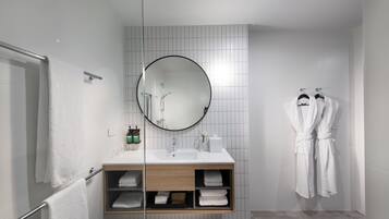 Un baño moderno con cabina de ducha de vidrio, un espejo redondo, un mueble de tocador de madera con lavabo y dos albornoces blancos colgados en ganchos.