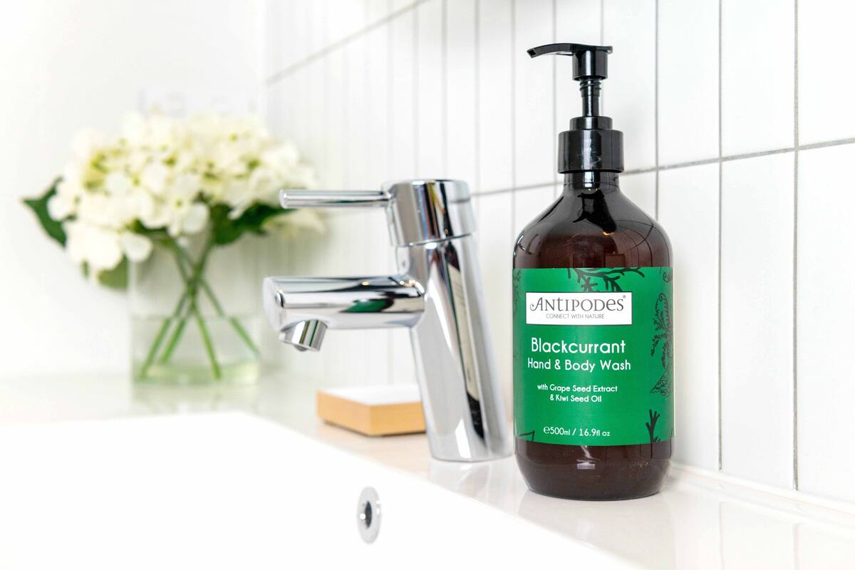 Une bouteille de gel lavant pour les mains et le corps à la cassis de la marque Antipodes, dotée d’un distributeur à pompe, posée sur une surface blanche à côté d’un robinet chromé.