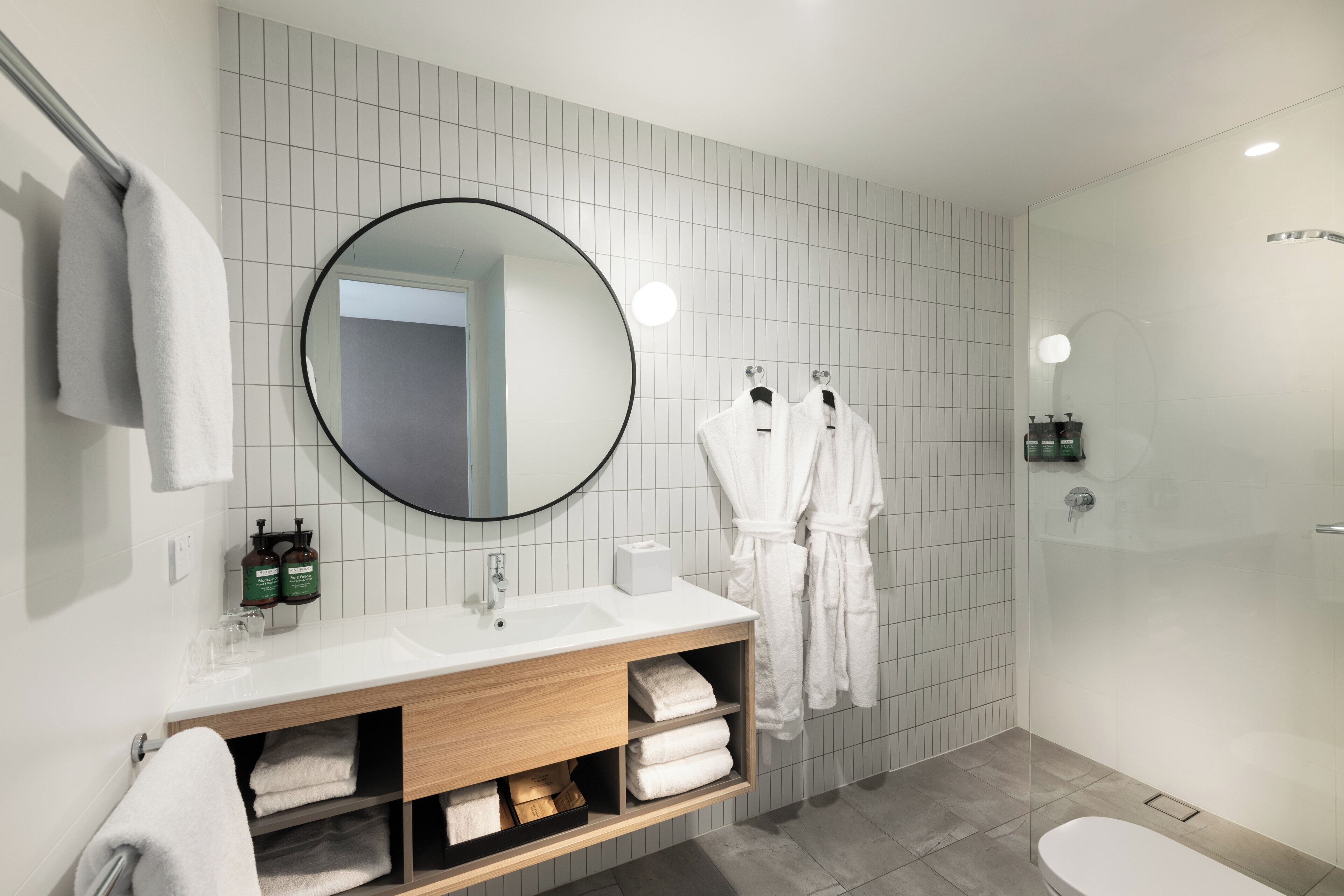 Un baño moderno con un espejo grande, azulejos blancos, un tocador de madera y una ducha con mampara de vidrio.