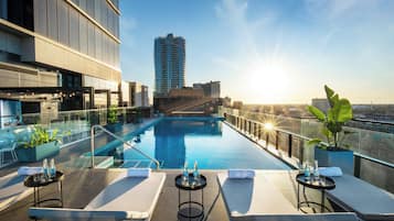 A rooftop pool with lounge chairs and tables, surrounded by glass railings and potted plants.