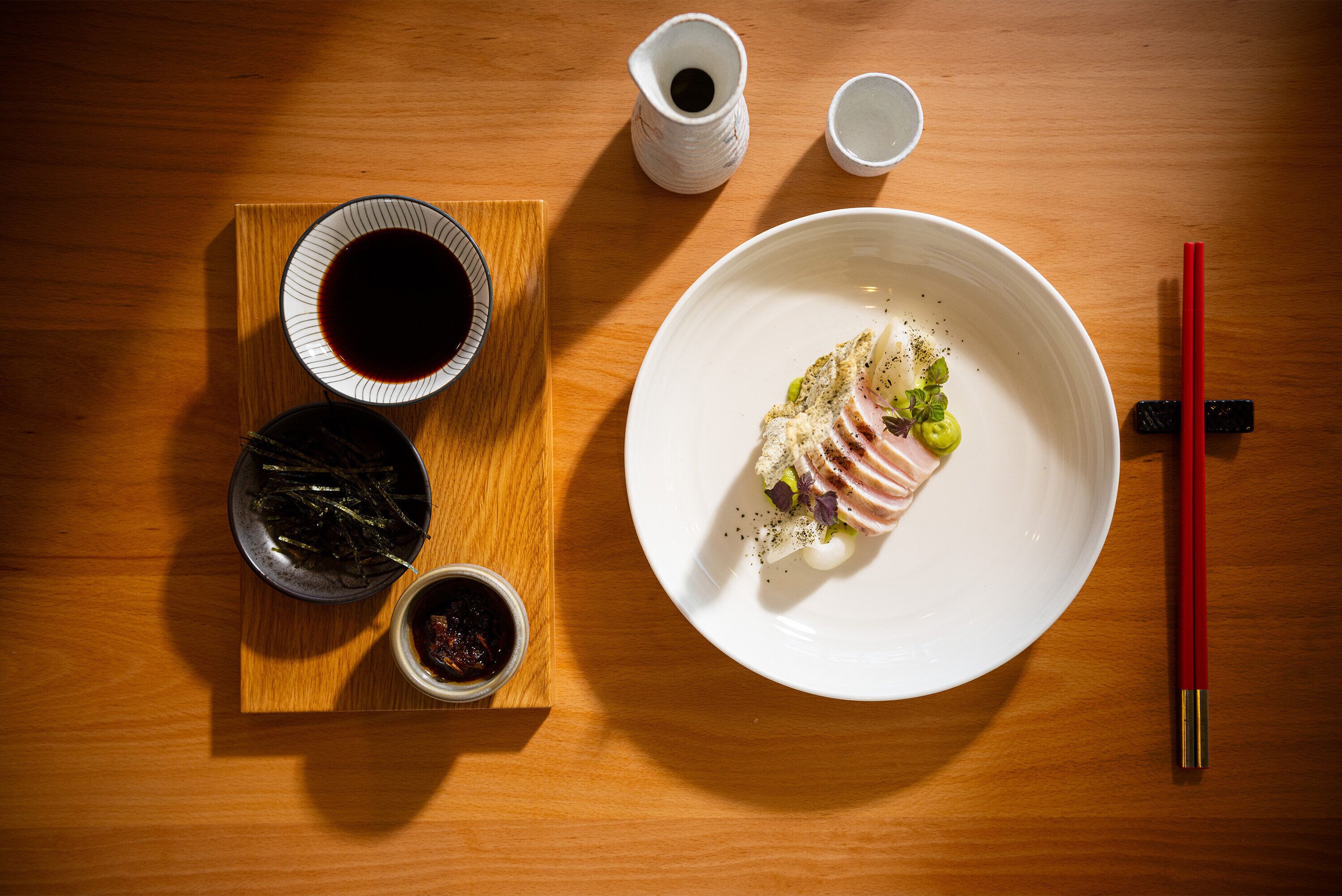 A wooden table with a white plate of food, two small cups of soy sauce, a bowl of pickled vegetables, and a pair of red chopsticks.