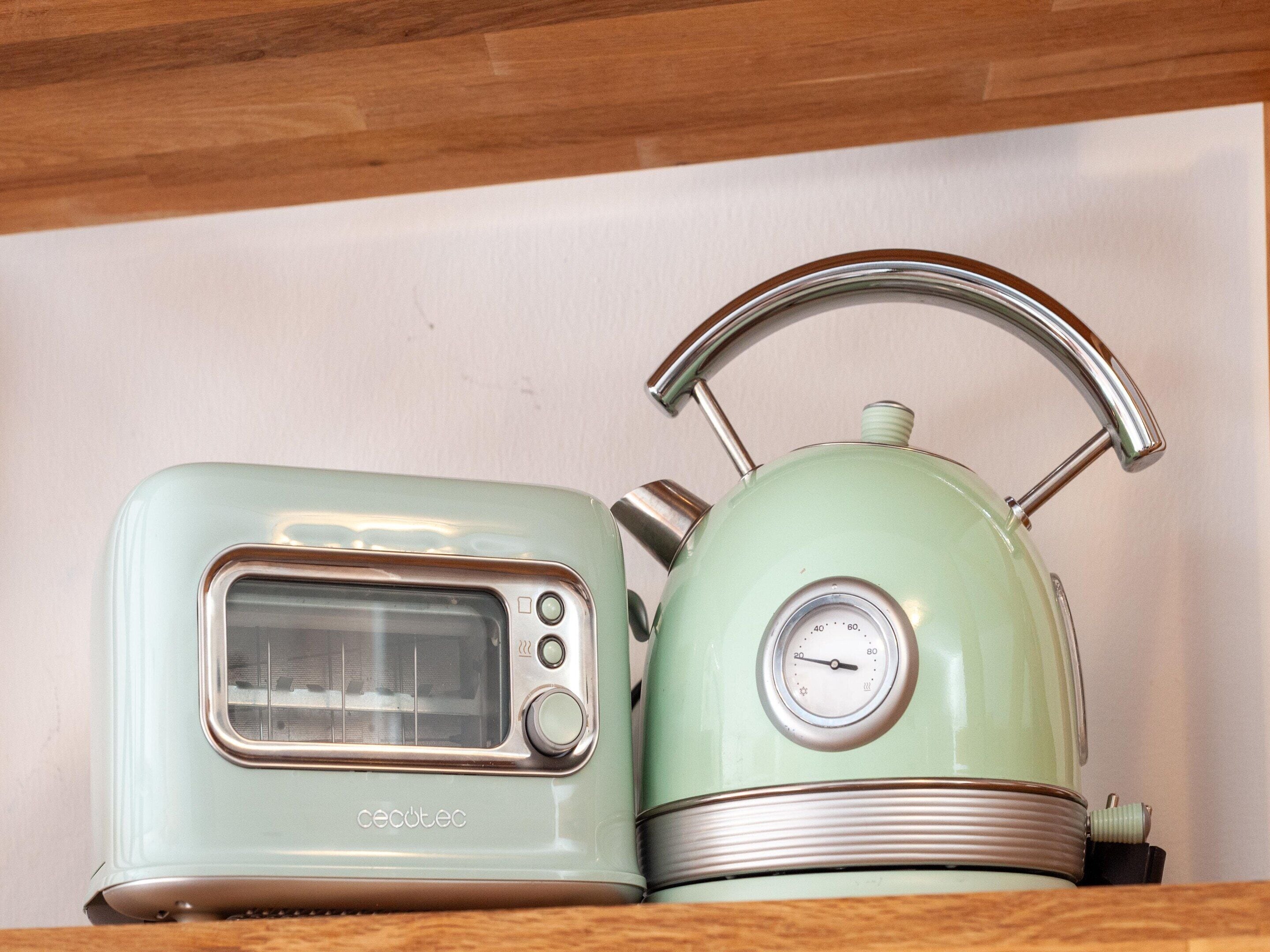 Un tostador y tetera vintage en color verde azulado, con asa metálica, sobre un estante de madera frente a una pared lisa.