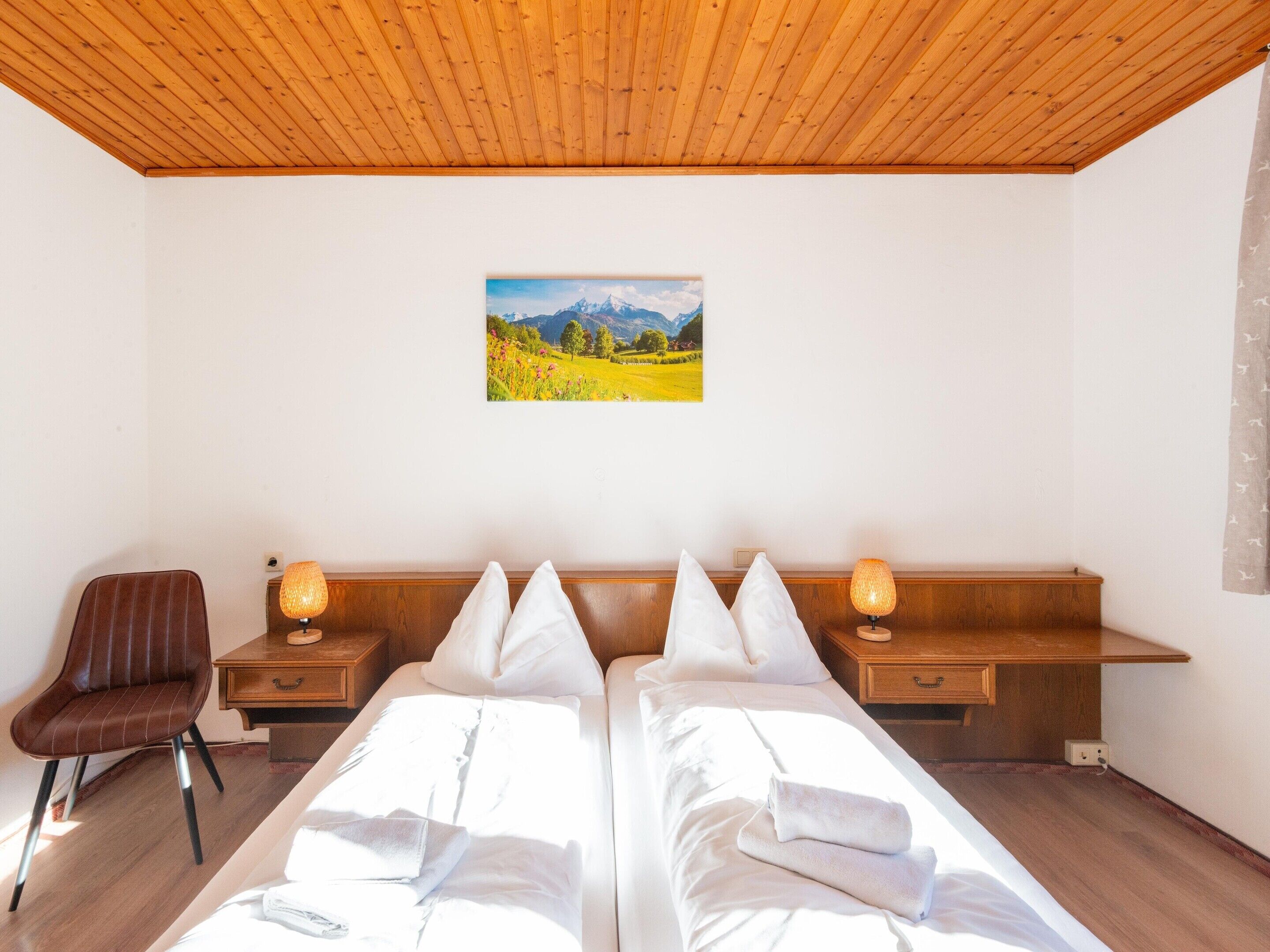 A hotel room with two beds, a wooden ceiling, a painting of a mountain landscape, a chair, and bedside lamps.