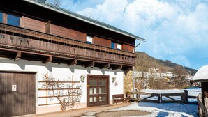 Ein traditionelles Holzhaus mit Balkon, Steinmauer und befestigtem Bereich mit Schneeflecken.