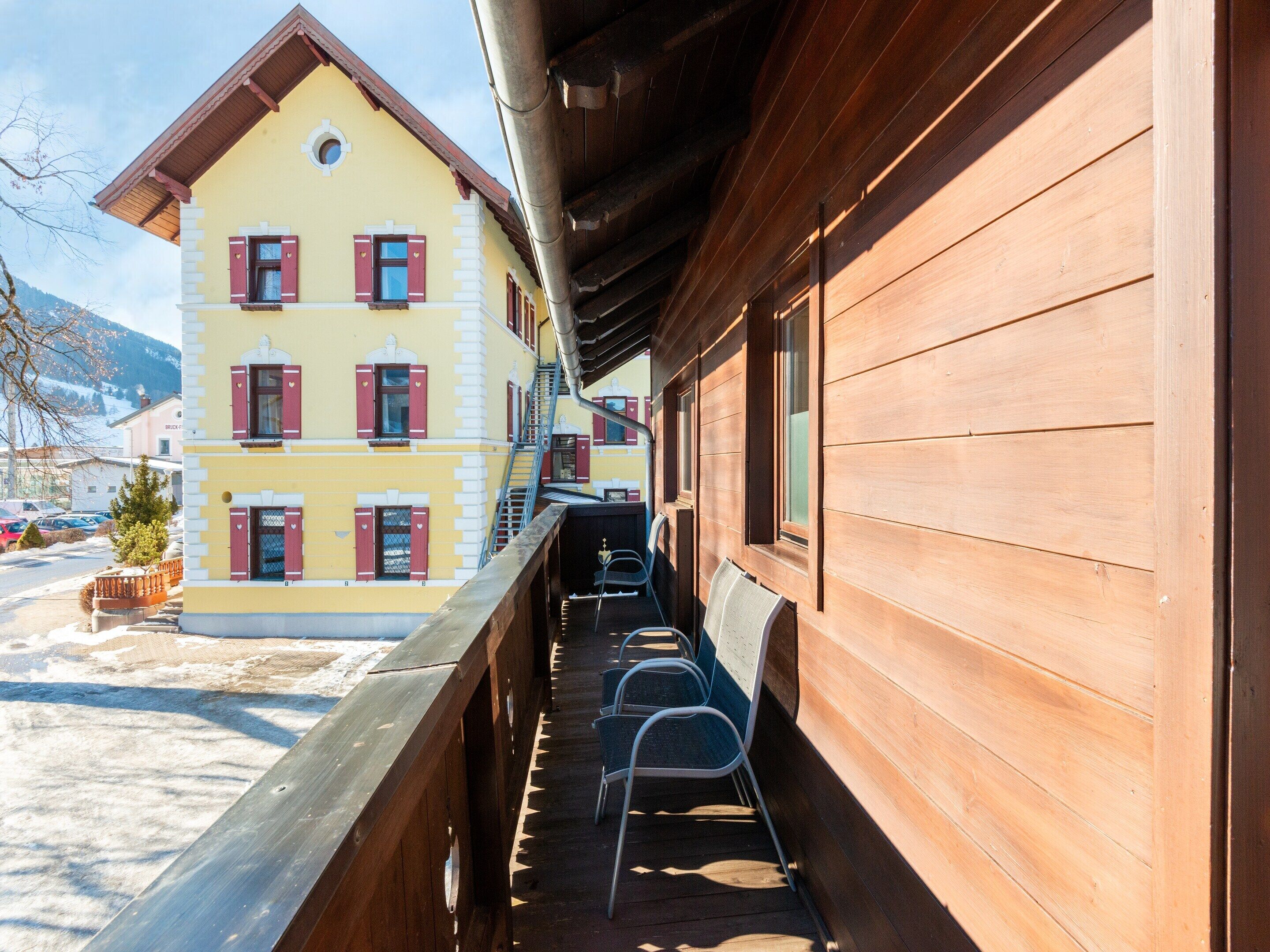 A wooden balcony with metal chairs, a wooden railing, and a yellow building with red shutters in the background.