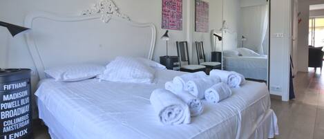 A neatly made bed with white linens and rolled towels, a large mirror, and a black trash bin with text on it.