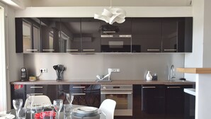 A modern kitchen with dark cabinetry, a glass dining table, and a hanging light fixture. - 1-Br Apartment, Sea View, Steps from Beaches (Cannes)