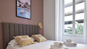 A neatly made bed with white and beige pillows, a grey headboard, and a window with a view of palm trees. - Modern 3-Br Apartment in City Center, 5 Minutes from Palais and Beache (Cannes)