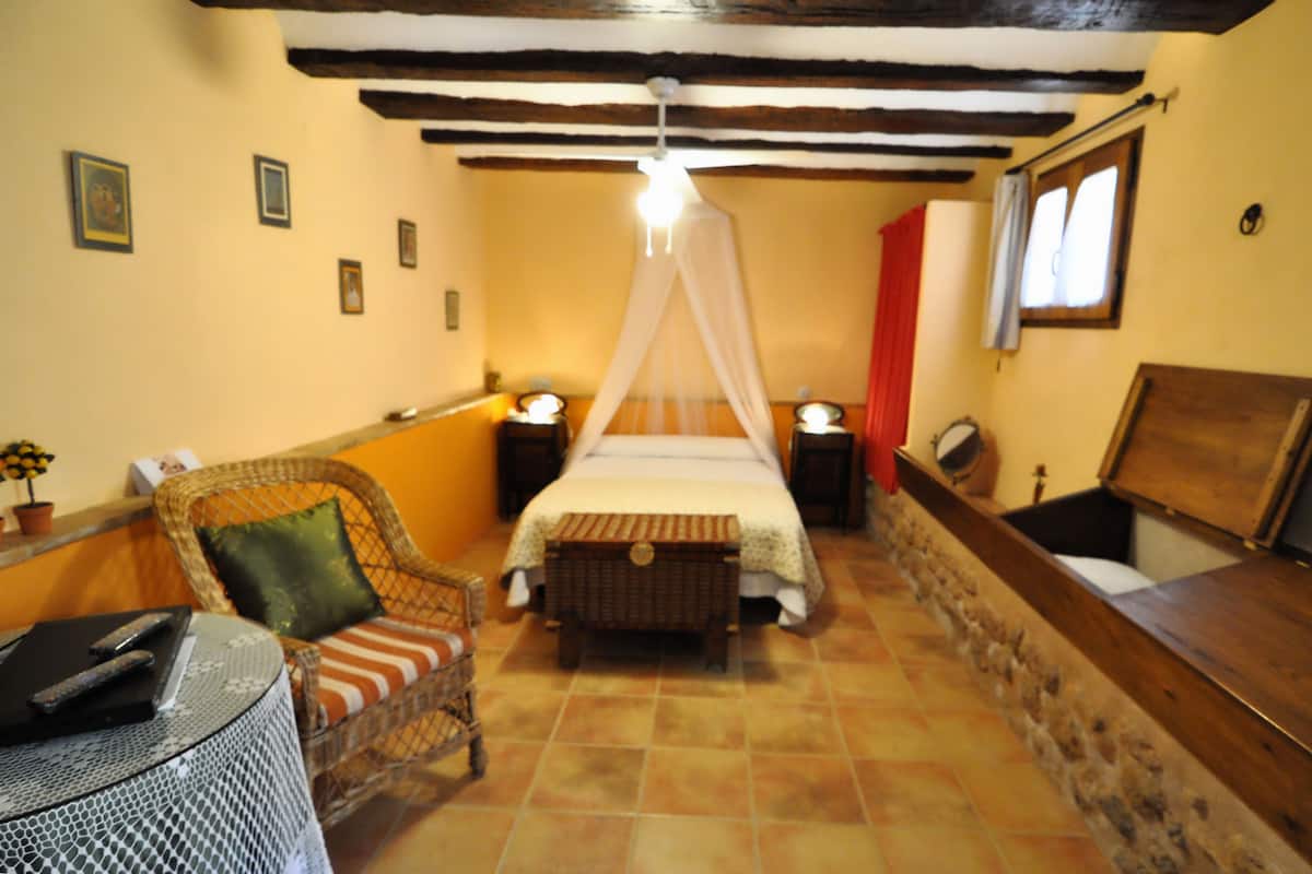Ein Schlafzimmer mit einem Himmelsbett, einem Korbstuhl, einem kleinen Tisch mit Fernbedienung und einer Holzbank.