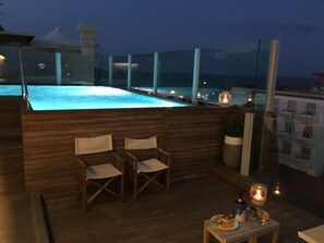 A rooftop pool area with wooden deck, two chairs, a small table with a candle, and a view of the cityscape at dusk. - Hotel City (Rimini)