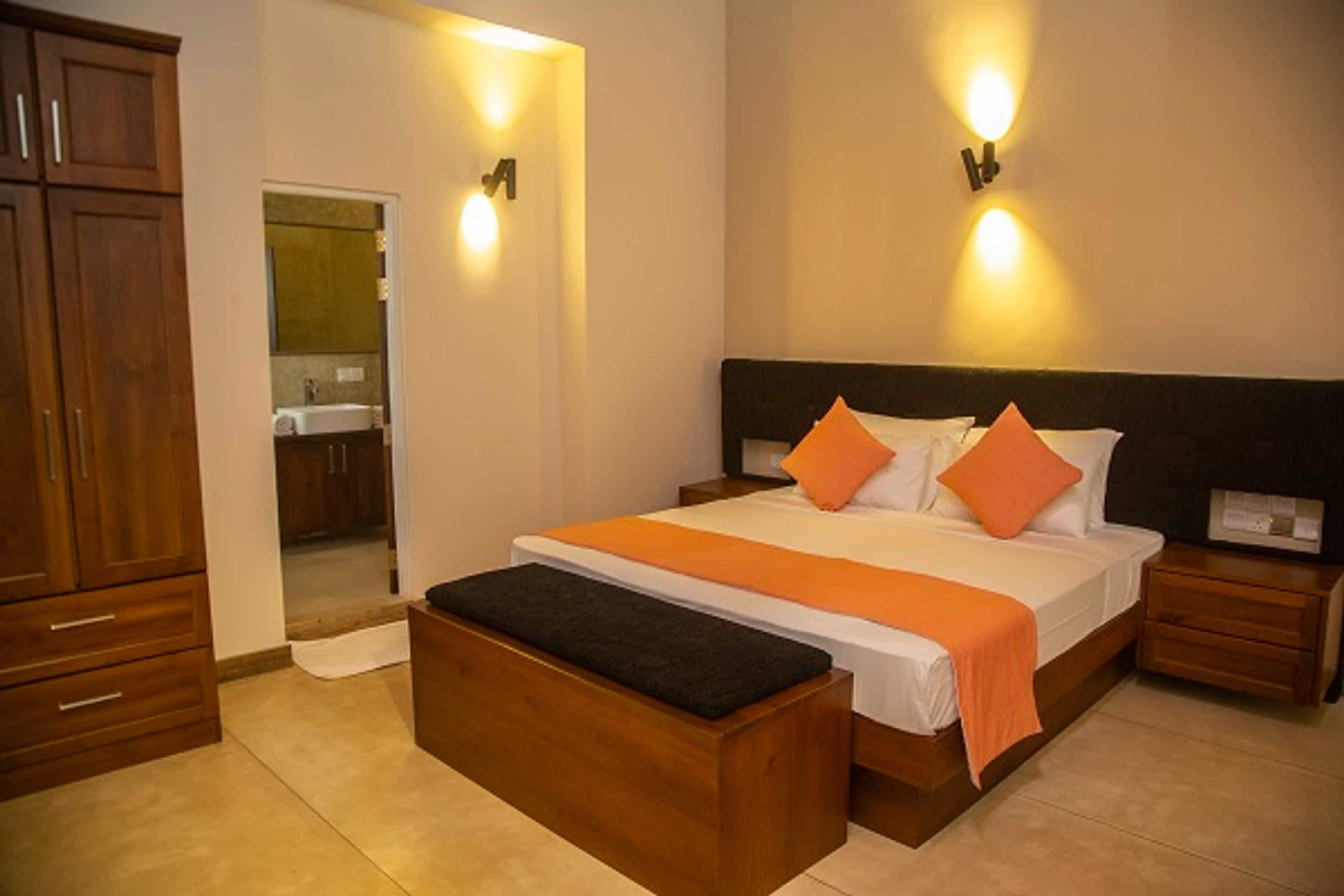 A bedroom with a bed, wooden nightstands, a bench, and a bathroom visible through an open door.