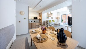 Un comedor moderno con una mesa de madera dispuesta para dos personas, una jarra negra y un florero con flores. Se ve una cocina y una sala de estar al fondo.