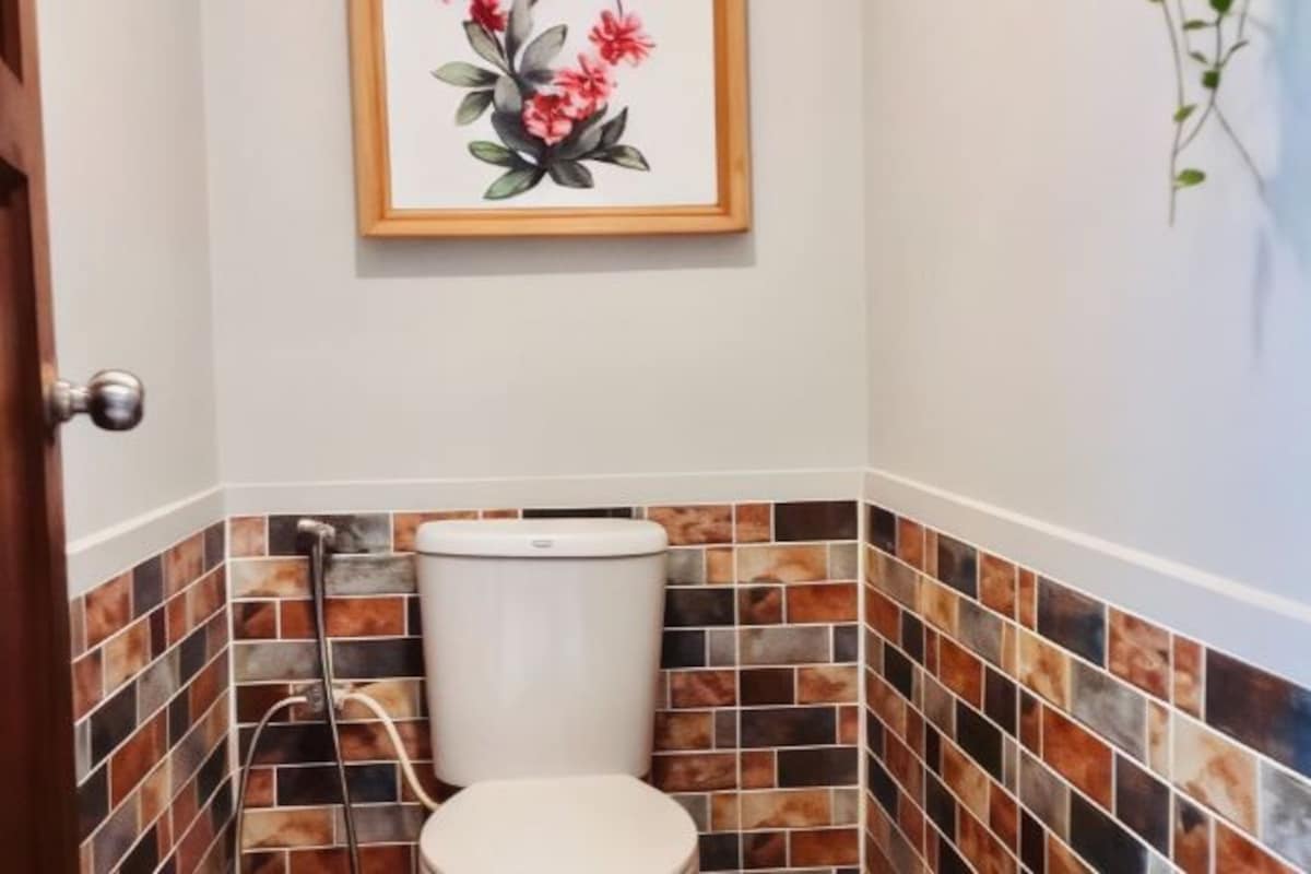 A bathroom with a toilet, a shower, and a tiled wall with a floral painting above the toilet.