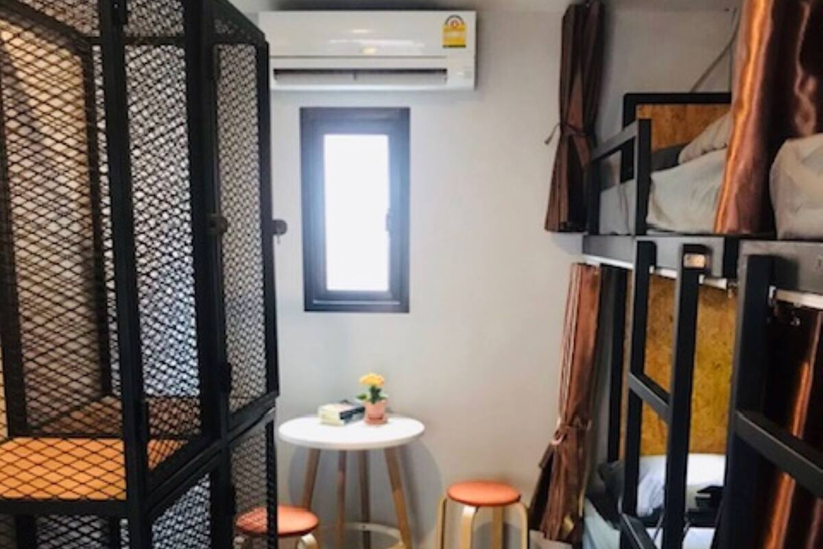 Ein kleines Zimmer mit Etagenbetten, einem runden Tisch mit Hocker und einer Klimaanlage an der Wand.