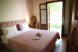 A neatly made bed with a pink bedspread and white pillows, a wooden nightstand with a lamp and a vase of flowers, and a door leading to a balcony. - Romantic ALOE villa in PRINES with private pool, sea-sunset view, near beach. (Rethymnon, Crete)