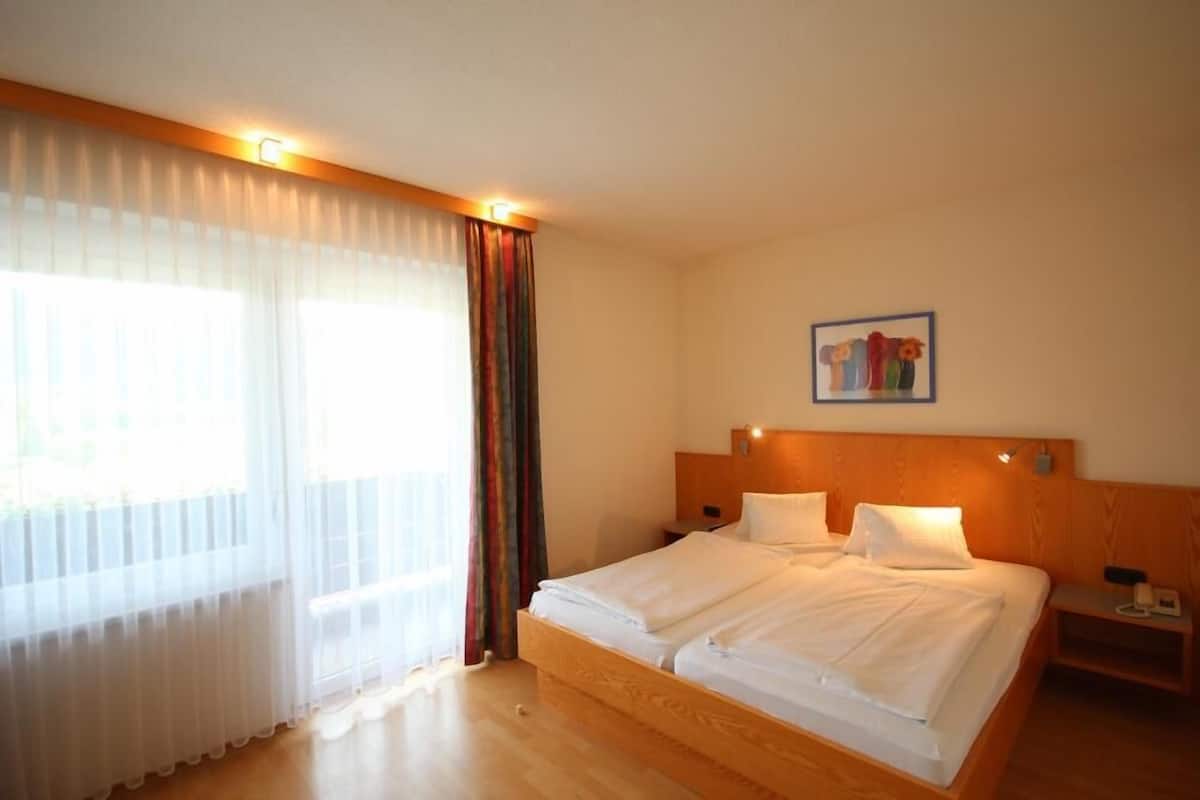 Une chambre d’hôtel avec un grand lit, une table de chevet, une lampe fixée au mur et une fenêtre avec des rideaux légers.