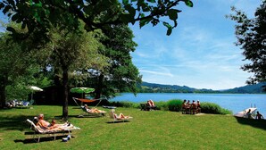 A grassy area with several lounge chairs and umbrellas, a dining table with people, and a view of a lake and hills in the background.