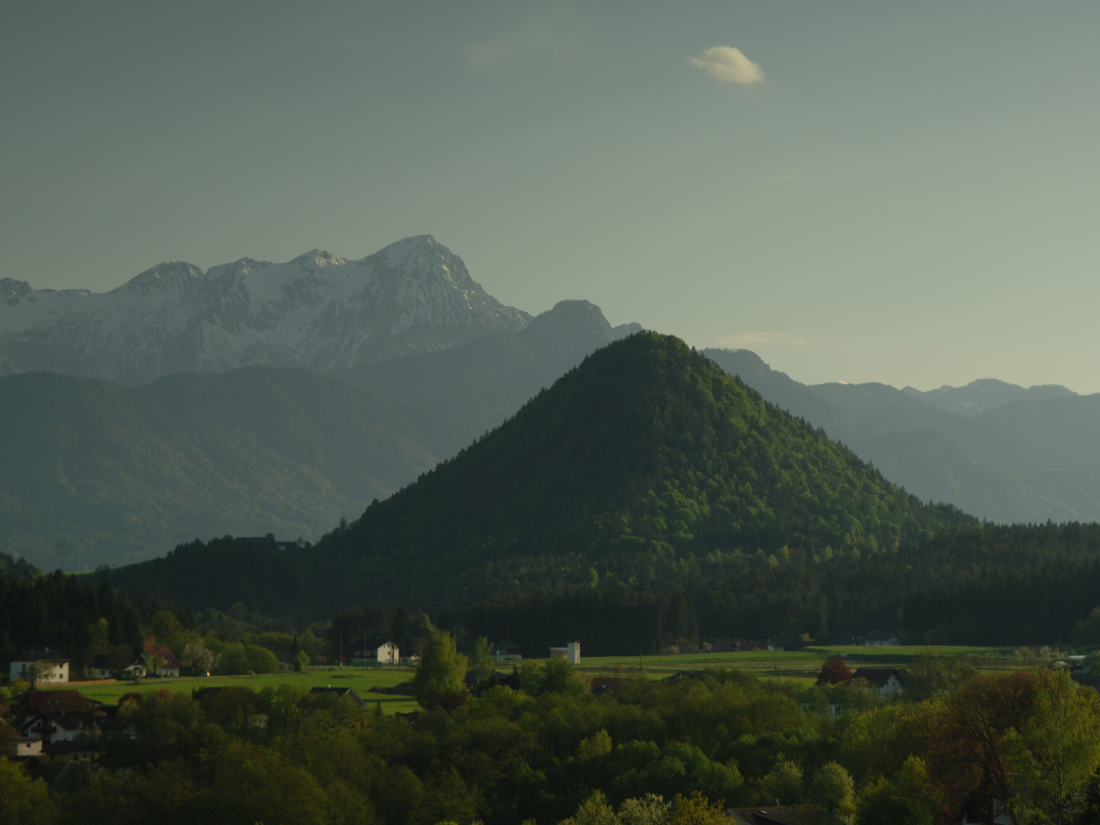 A mountainous landscape with a prominent hill in the foreground and a range of mountains in the background. There are buildings and greenery in the valley below.