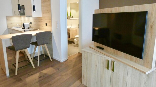 Eine moderne Küche mit Essbereich, einem großen Flachbildfernseher an einer Holzwand und einem sichtbaren Badezimmer im Hintergrund.