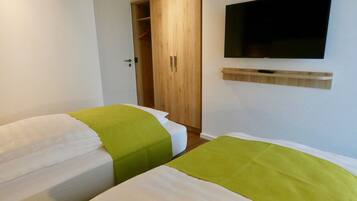 A hotel room with two beds, a wooden wardrobe, a television mounted on the wall, and a shelf below it.