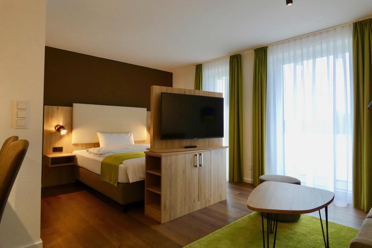 A hotel room with a bed, a TV mounted on a wooden cabinet, a small table, and a green rug.