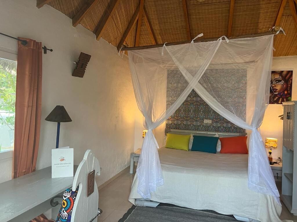 A room with a bed under a mosquito net, a bedside table, a lamp, and a wall with a patterned design.