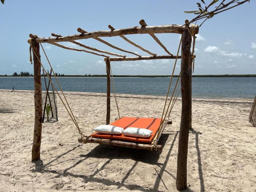 En gyngestol i træ med madras og puder, placeret på en sandstrand med et vandareal i baggrunden.