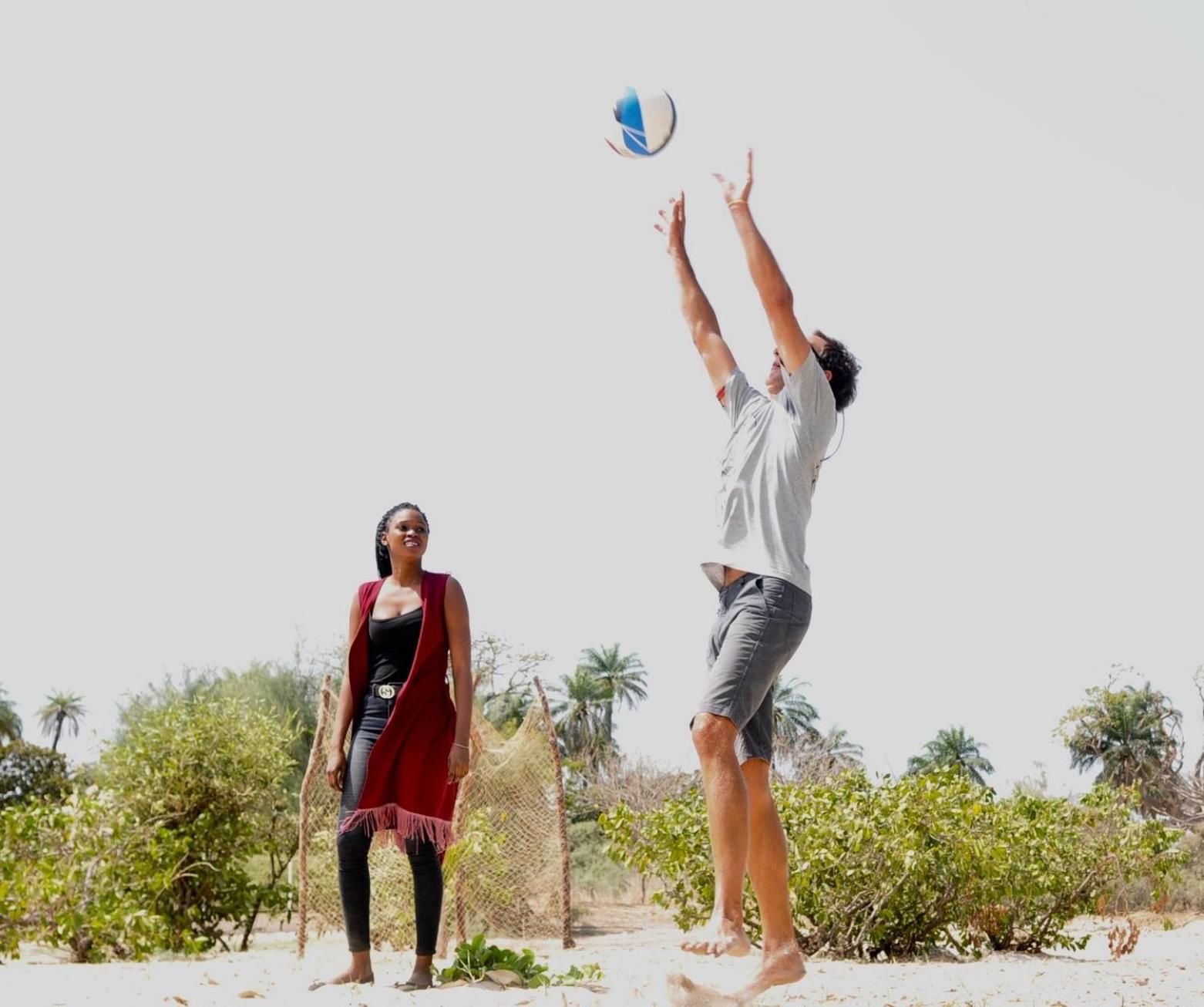 Two people playing beach volleyball on a sandy beach with sparse vegetation and a clear sky.