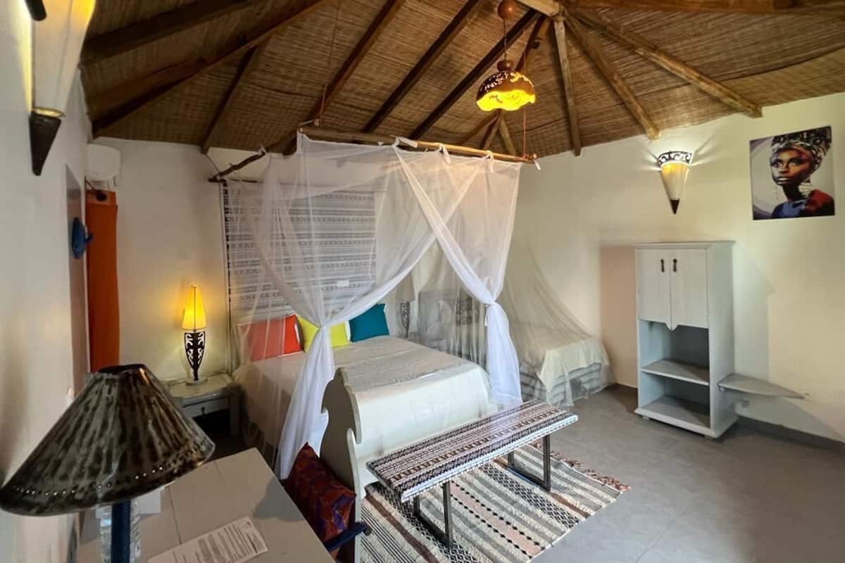 Une chambre avec un lit sous un toit de chaume, une lampe de chevet, une petite table, une armoire et un luminaire fixé au mur.