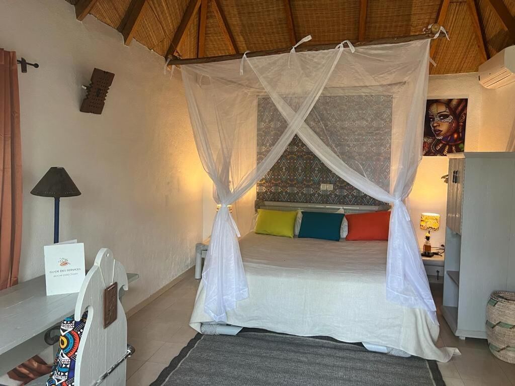 A room with a bed under a mosquito net, a bedside table, a lamp, and a wall with a patterned design.