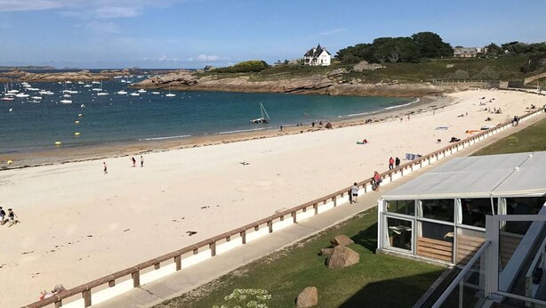 A beach with a clear view of the sea, a sandy shore, and a few boats. There is a building with a glass facade in the foreground.