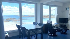A room with a dining table, chairs, a TV, and large windows with a view of the sea.