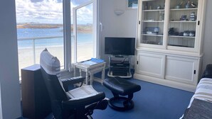 A room with a large window overlooking a beach, a television on a stand, a cabinet with kitchenware, and a massage chair.