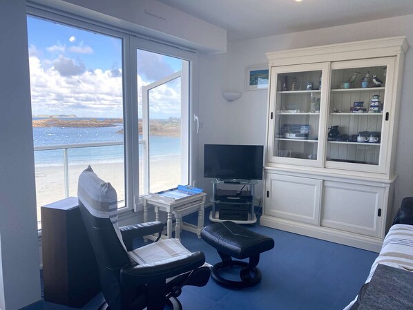 A room with a large window overlooking a beach, a television on a stand, a cabinet with kitchenware, and a massage chair.