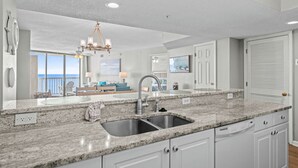 A modern kitchen with granite countertops, a double sink, white cabinetry, and a view of the ocean through large windows.