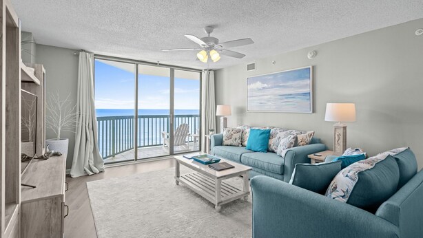 A spacious living room with a large sliding glass door leading to a balcony with a view of the ocean. There is a ceiling fan, a coffee table with books, and a blue sofa with patterned cushions.