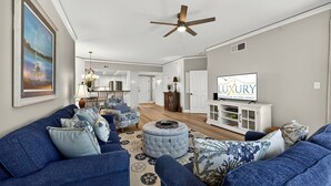 A spacious living room with blue sofas, a round ottoman, and a television displaying 'LUXURY RENTALS OF HILTON HEAD'.