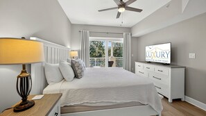 Une chambre à coucher comprenant un lit, une table de chevet, une lampe, un ventilateur de plafond, un téléviseur et un balcon avec des rideaux.
