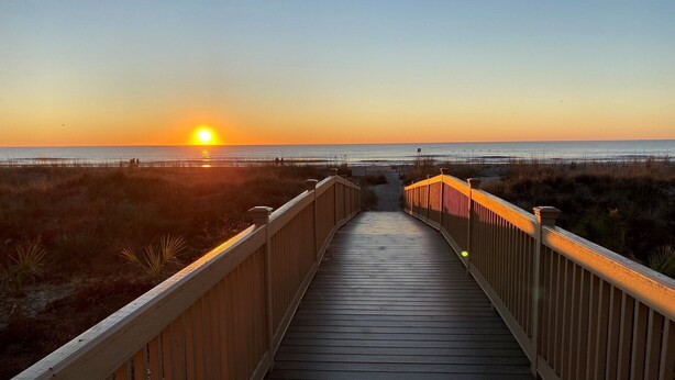 A wooden boardwalk leading to a beach with the sun setting in the background.