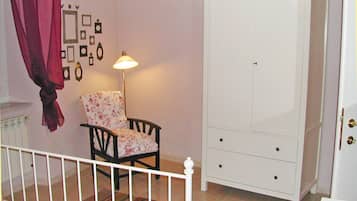 A bedroom with a white wardrobe, a floral chair, a bedside table, and a lamp.