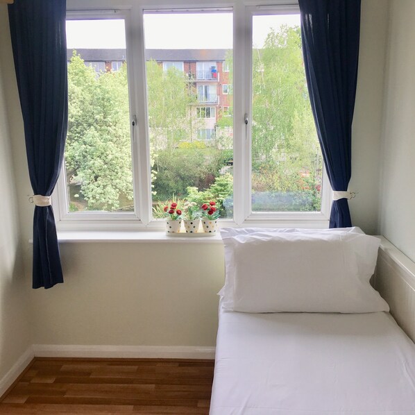 A bedroom with a bed, a window with curtains, and a view of a residential area with trees and buildings. - Apple Perch 2BR Flat-Free Parking (Oxford)