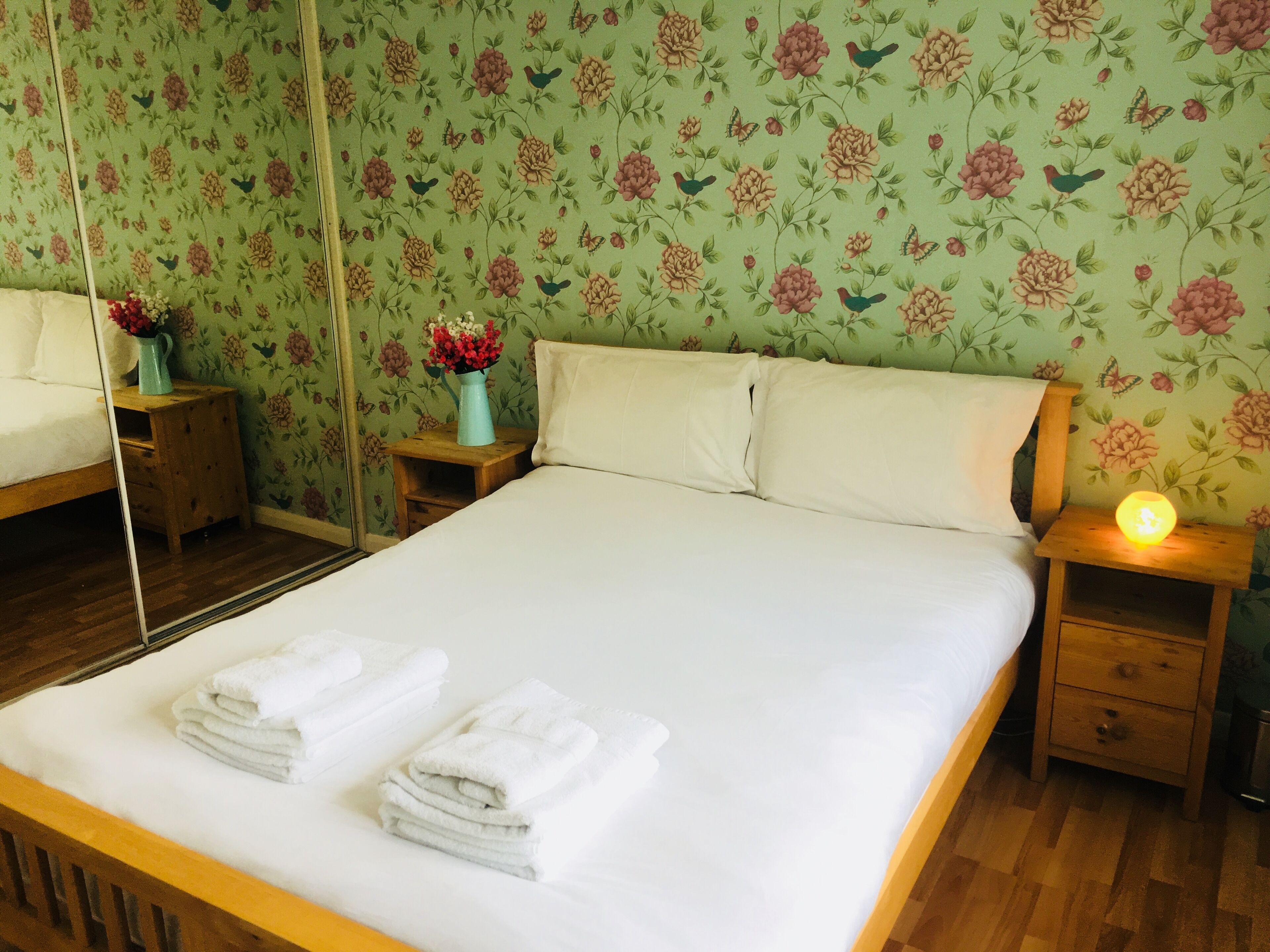 A neatly made bed with white linens and towels, a wooden nightstand with a vase of flowers, and a bedside lamp.