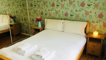 A neatly made bed with white linens and towels, a wooden nightstand with a vase of flowers, and a bedside lamp.