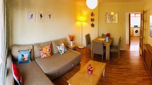 A living room with a sofa, coffee table, and dining area. There is a TV, a lamp, and a washing machine visible in the background. - Apple Perch 2BR Flat-Free Parking (Oxford)