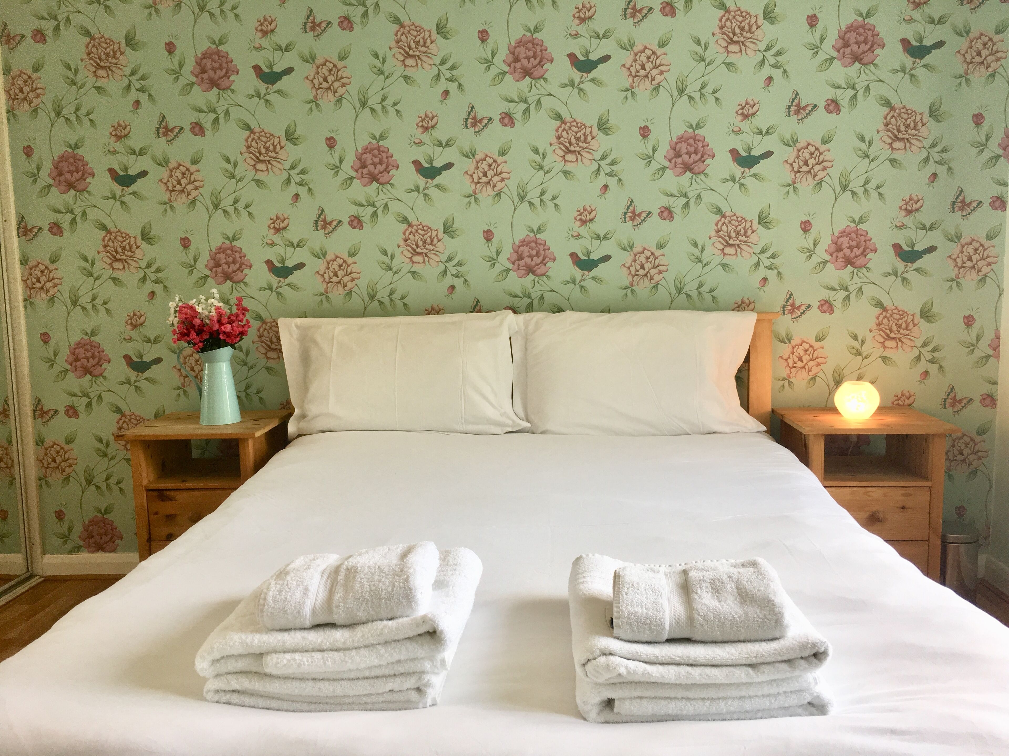 A neatly made bed with two folded towels, a vase with flowers, and a lit bedside lamp.