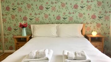 A neatly made bed with two folded towels, a vase with flowers, and a lit bedside lamp.