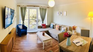 A living room with a sofa, a blue armchair, a coffee table, a TV mounted on the wall, and a dining table with a vase of flowers. - Apple Perch 2BR Flat-Free Parking (Oxford)