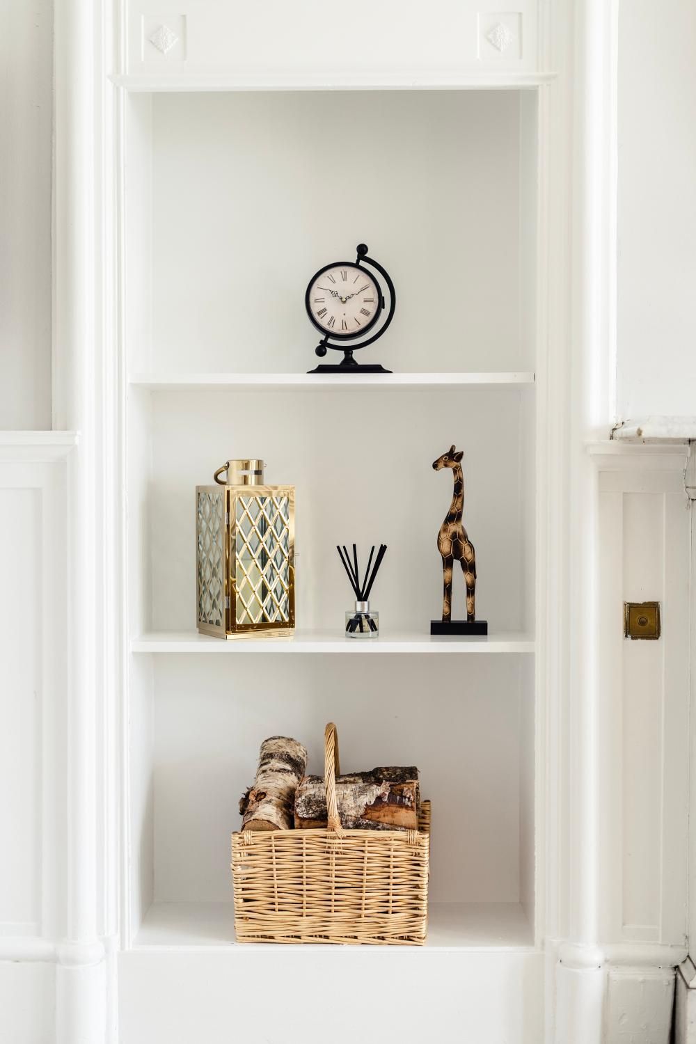 A white shelf with a clock, a decorative box, a reed diffuser, a giraffe figurine, and a wicker basket.