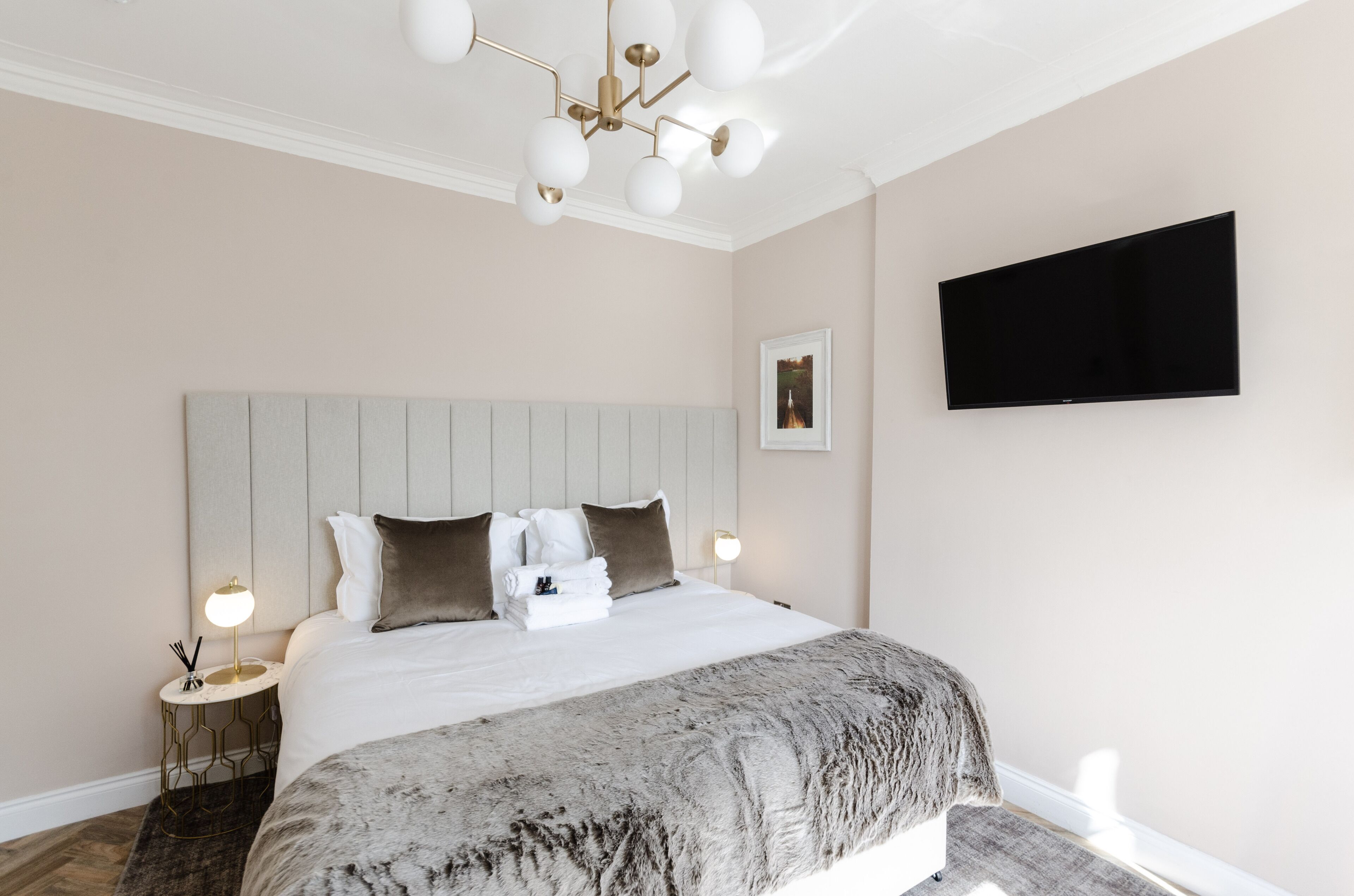A neatly made bed with a grey blanket, a wall-mounted TV, and a modern chandelier.